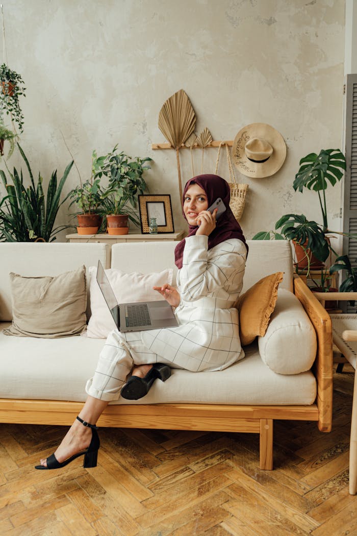 A smiling Muslim woman in hijab working on a laptop while making a phone call, sitting on a stylish sofa indoors.